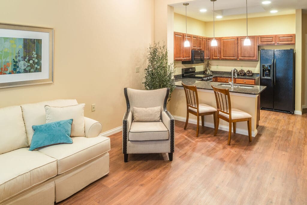 A living room area with a beige couch, beige sofa chairs, with access to the kitchen with light brown cupboards, gray marbled counters, bar stools, a sink, and a black fridge.