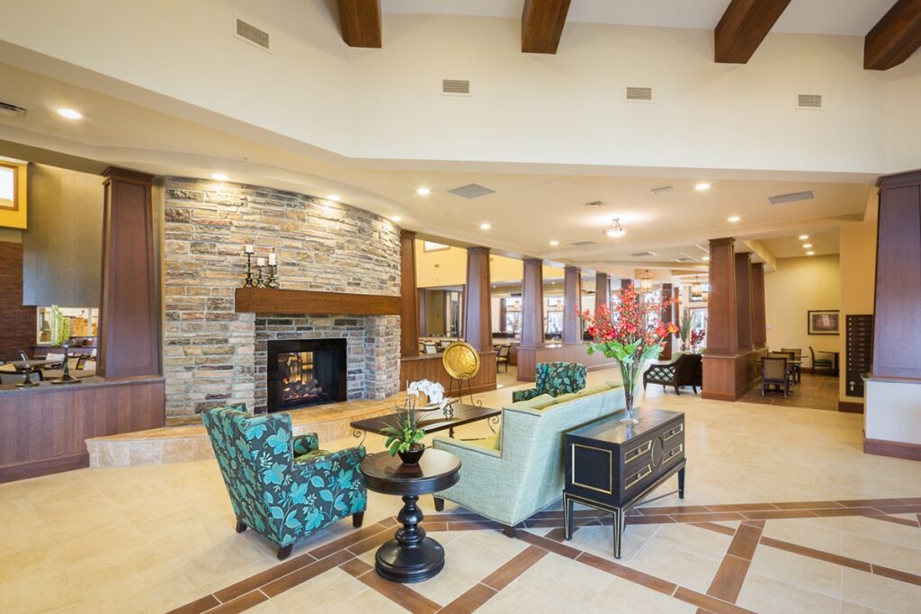 The grand lobby area with a light blue couch, blue sofa chairs, side tables with vases of flowers on them, all in front of a fireplace at Sumter Senior Living.