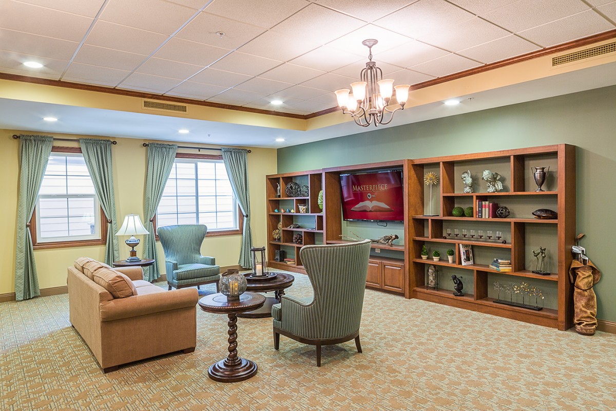 Grand neighborhood first floor sitting area with a light brown couch, light blue sofa chairs, side tables with lamps on them, and a wall-length shelf and a TV.