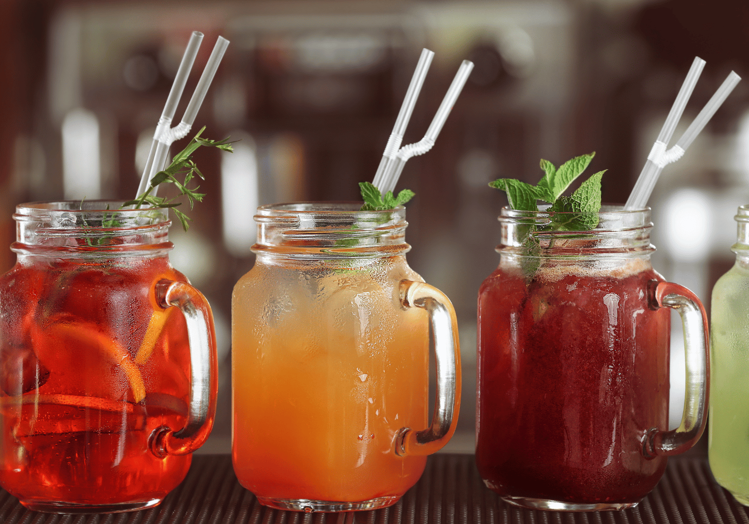 Three mason jar cocktails with straws and mint garnish on a bar, from red to orange to yellow beverages.