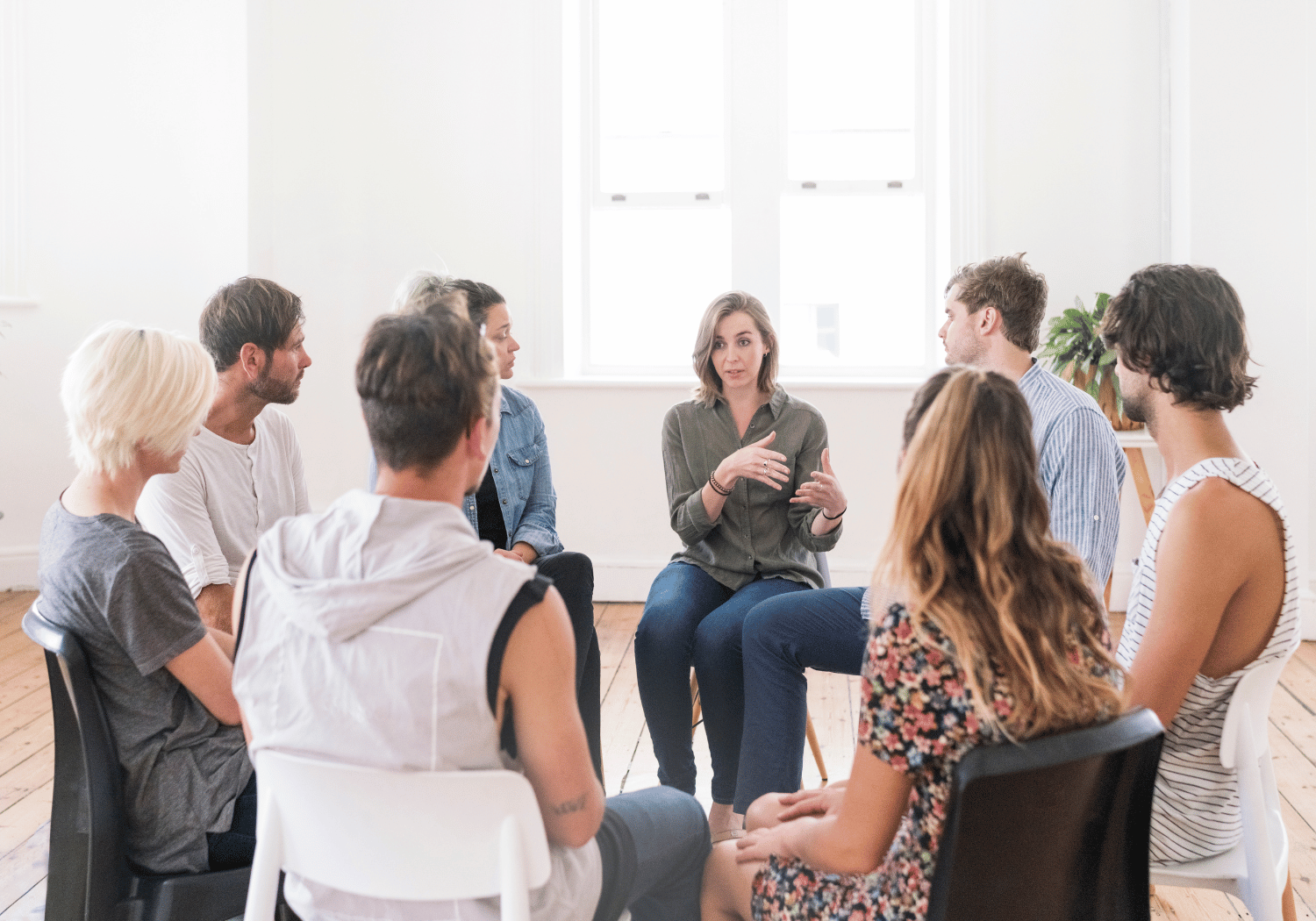 Group of adults seated in a circle in a bright room; a woman in an olive blouse speaks and gestures.